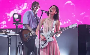 A female singer with a glittering guitar sings into a microphone while a male guitarist plays in the background.