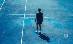 A tennis player stands on a blue court, facing away from the camera, holding a racket.
