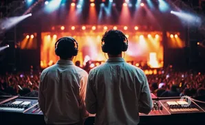Two DJs in headphones stand facing a bright stage and cheering crowd at a concert.