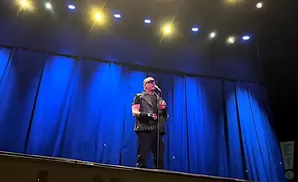 A bald man in sunglasses and a vest sings into a microphone on a stage with blue curtains.