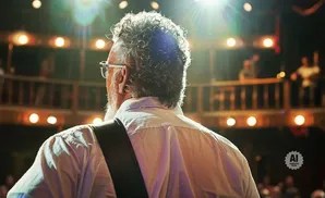 An older man with curly hair and glasses stands with his back to the camera, facing a bright stage with blurred lights and people.