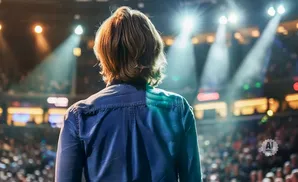 A person from behind in a blue shirt stands on a stage facing a bright spotlight and a large audience.