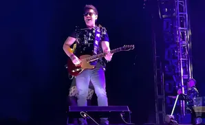 A man in sunglasses plays a red electric guitar on a stage with purple lighting and a metal scaffolding structure.