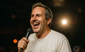 A man laughs into a microphone, wearing a white t-shirt, with a blurred light behind him.