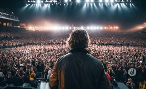 A performer facing a huge, cheering crowd at a concert, with bright stage lights overhead.
