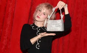 A woman with pink hair and star makeup holds a pearl beaded handbag in front of a red velvet curtain.
