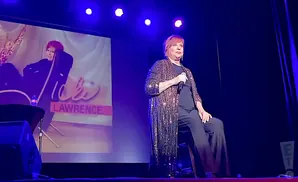 A woman with red hair and a sparkling robe is sitting on a stage, holding a microphone.