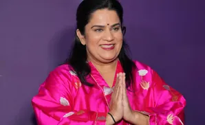 A woman in a pink robe with embroidered flowers smiles and places her hands together in a greeting.