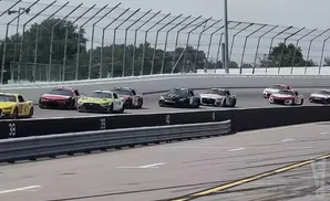 A pack of NASCAR race cars in formation on a track with barriers and fencing.