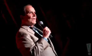 A man in a light-colored suit and blue shirt holds a microphone, looking to his left.