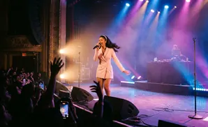 Singer performs on stage in a pink blazer dress as a DJ plays in the background with purple and blue stage lights.