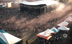 Aerial view of a large outdoor concert crowd with a stage and tents, bathed in sunlight and dust.