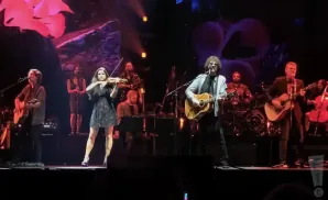 A band performs on stage, with a violinist in the foreground and guitarists and a drummer behind her.