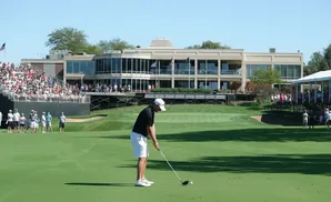 Golfer in black shirt and white shorts prepares to swing on a sunny golf course with a large building and crowd in the background.