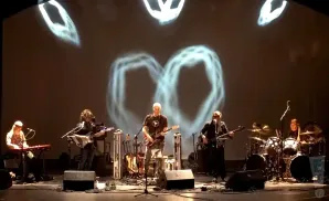 Musicians perform on stage with abstract visuals projected behind them.