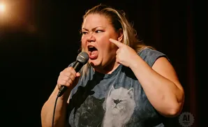 A woman with blonde hair on a stage, pointing at her cheek while holding a microphone.
