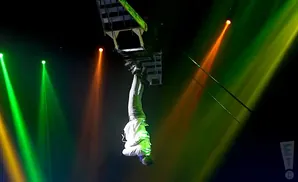 A performer hangs upside down from a trapeze, illuminated by green and orange stage lights.