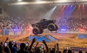 Monster truck jumps over a dirt mound as a large crowd watches with their phones out.