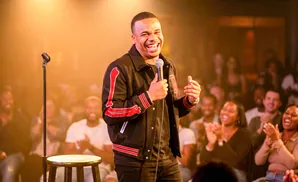 A comedian laughs on stage, holding a microphone, with a laughing audience in the background.