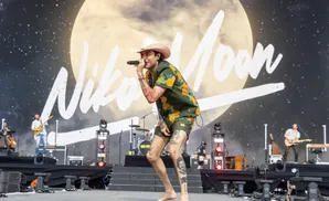 A male singer in a cowboy hat and orange fruit-print shirt performs on stage, holding a microphone.