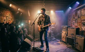 A male guitarist performs on stage in front of a brick wall plastered with posters, while the audience watches.