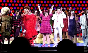 A cast in colorful costumes on stage, with a bright red and purple dotted backdrop.