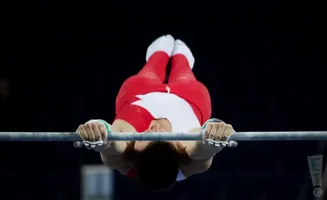 Gymnast in red and white suit hangs upside down from a bar, gripping it with chalked hands.