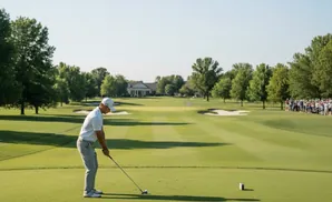 Golfer preparing to tee off on a sunny golf course with a clubhouse and spectators in the distance.