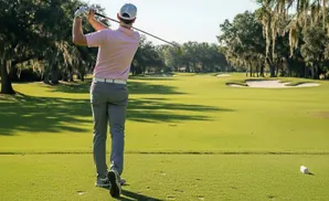 Golfer in pink shirt and grey pants swings club on a green golf course with sand traps and trees.