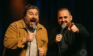 Two men are on stage with microphones, laughing. One man wears a brown jacket, the other a black jacket.