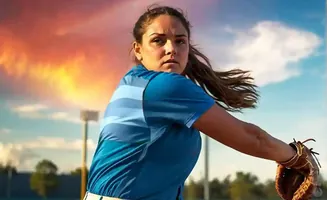 A softball pitcher winds up to throw, with a dramatic sunset sky in the background.