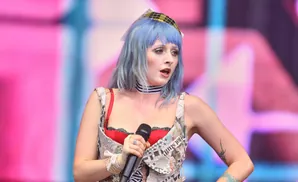 Singer with blue hair and newspaper outfit performing on stage with a pink and blue background.
