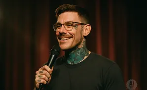 Man with glasses and tattoos smiles while holding a microphone in front of a dark curtain.