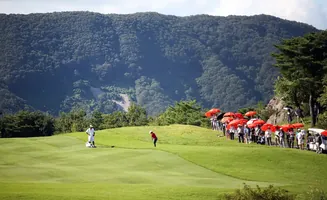 A golfer swings on a lush green course, with spectators holding orange umbrellas in the background.