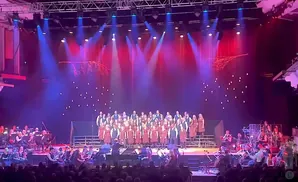 A choir and orchestra perform on a stage with red and blue lighting and decorative branches.
