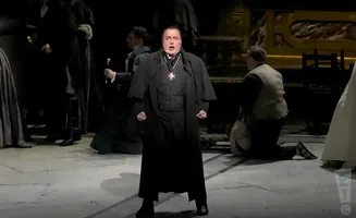 Opera singer in a black robe and medal sings with fists clenched.