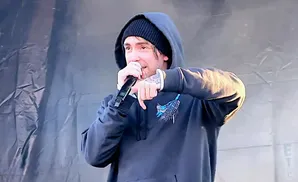 A man in a black beanie and hoodie holds a microphone and gestures with his hand.