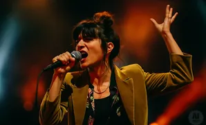 Woman with hair in a bun sings into a microphone on stage, wearing a gold velvet jacket.