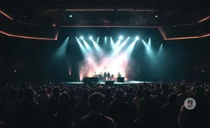 A band performs on a stage with bright lights and fog to a cheering crowd in a concert hall.