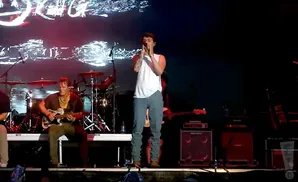 A male singer in a white shirt and patterned pants performs on stage with a guitarist and drums in the background.