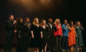 A diverse group of ten singers stand on stage holding microphones and smiling.
