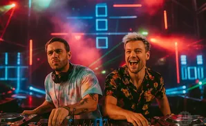 Two DJs at turntables with colorful lights and smoke in the background.
