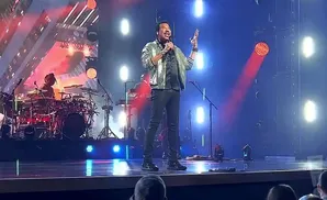 Lionel Richie on stage in a sparkly jacket, singing into a microphone with a band behind him.
