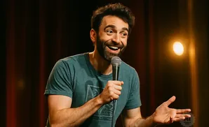 A man with a beard tells a joke into a microphone on stage.