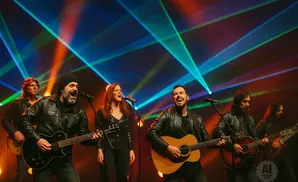A band performs with guitars and microphones under colorful laser lights.