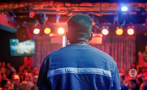 Man wearing a blue sweatshirt with a white stripe, seen from behind, facing a stage with red and orange lights.
