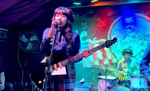 A female bassist sings into a microphone with a guitar. A drummer plays drums in the background.