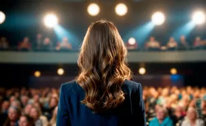 A woman with long, wavy brown hair stands with her back to the camera on a stage, facing a blurred audience.