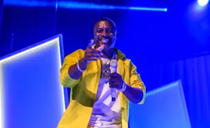 Akon in a yellow jacket smiles and points while holding a microphone.
