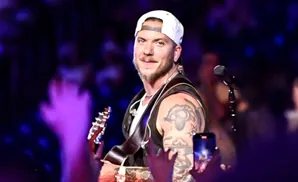 Man with tattoos and a white baseball cap plays guitar on stage with purple lights.